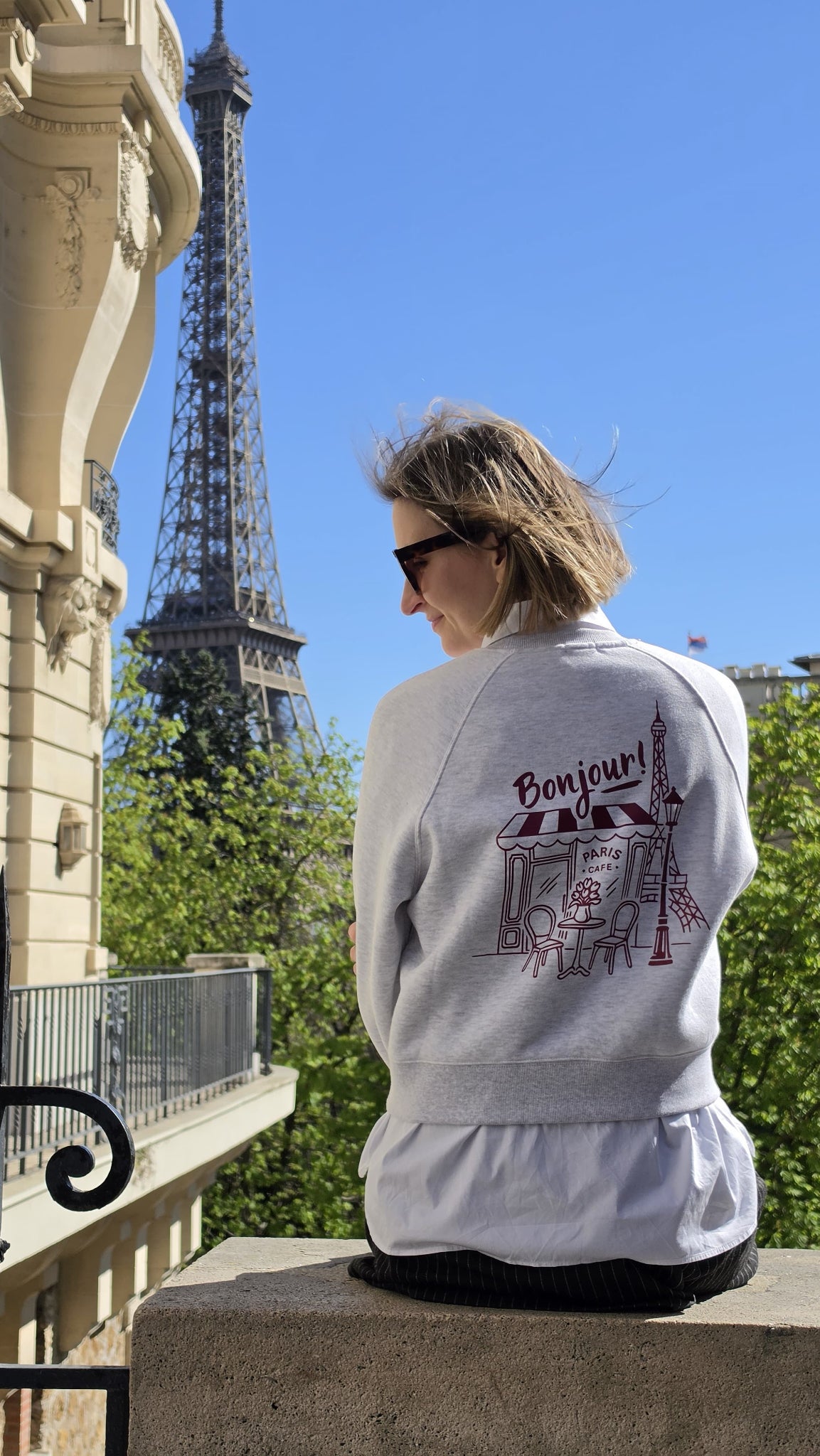 Anett vorm Eiffelturm in ihrem hellgrauen Sweater und einem großen "Paris Vibes" Backprint in bordeaux.