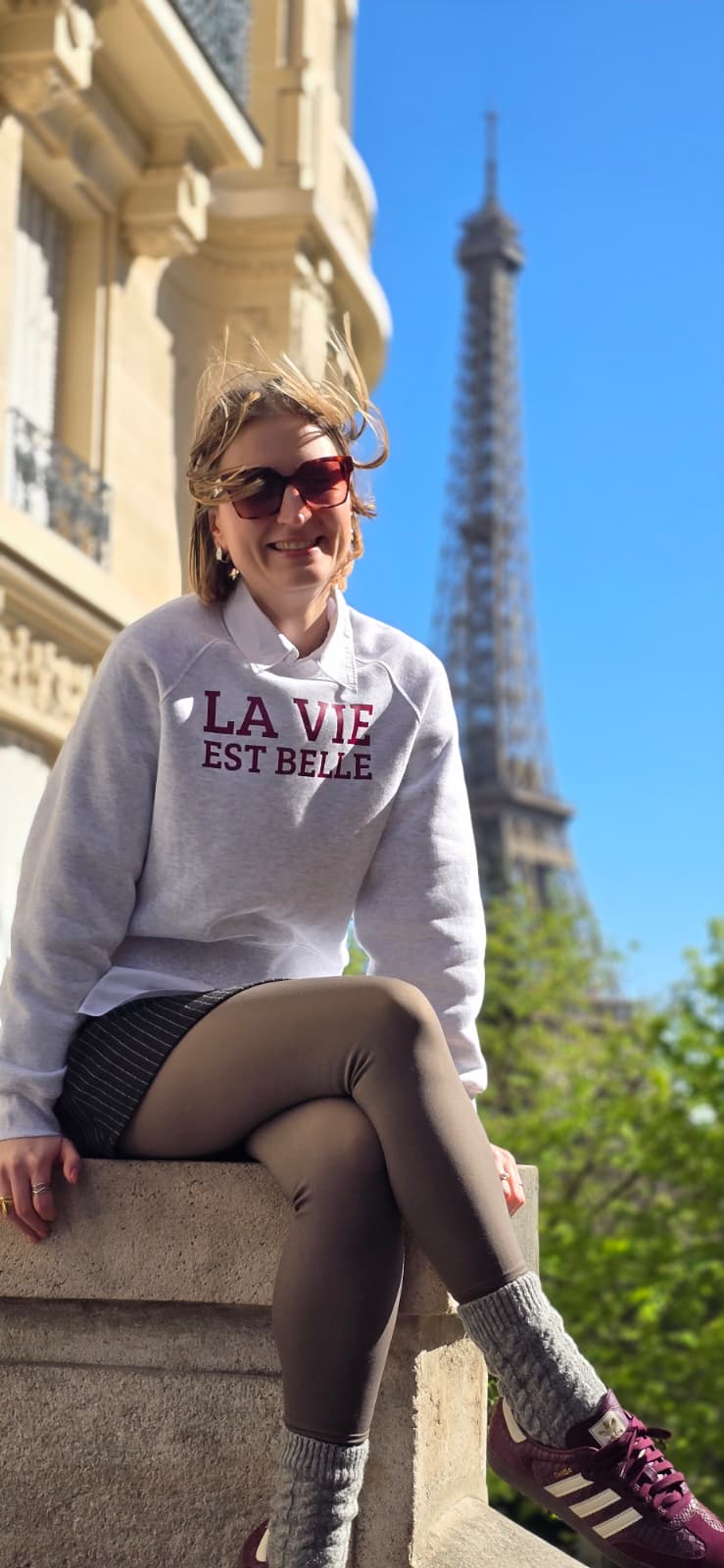 Anett vorm Eiffelturm in ihrem hellgrauen Sweater und einem großen "Paris Vibes" Frontprint in bordeaux.
